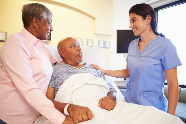 Photo of patient in bed, their hand being held by their partner. A clinician speaks to them