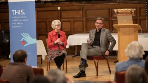 Mary Dixon-Woods (left), Director of THIS Institute, and Niles Peek (right), Professor of Data Science and Healthcare Improvement, sit on chairs at the front of the Great Hall at Homerton College, holding microphones and smiling while addressing an audience during Q&A.